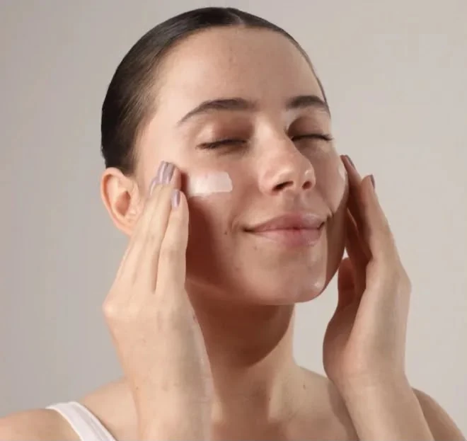 Mulher aplicando o Protetor Clareador Hidrabene no rosto, com os olhos fechados.